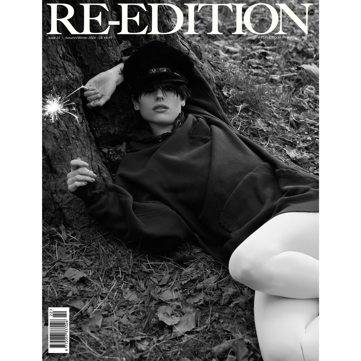 Monochrome magazine cover shot of a person in a hoodie and cap reclining on leaves next to a tree holding a sparkler.