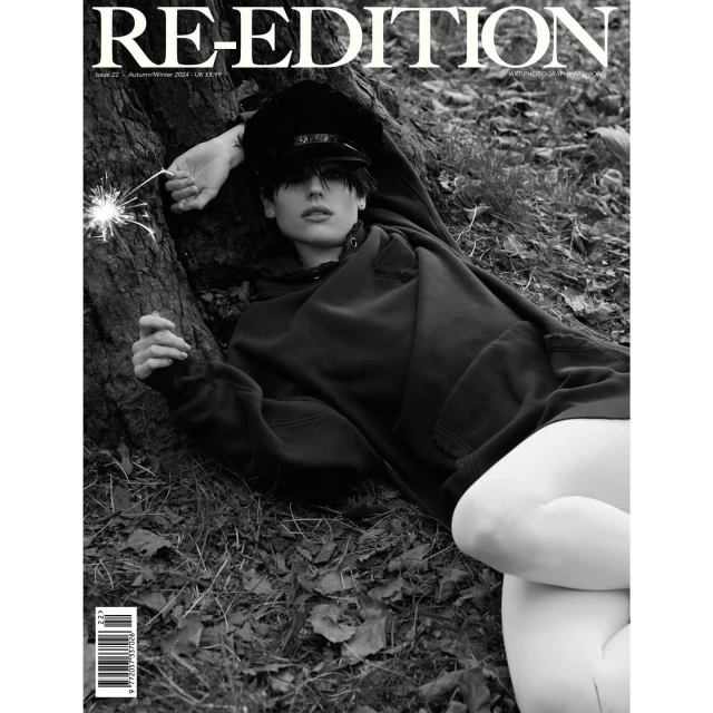 Monochrome magazine cover shot of a person in a hoodie and cap reclining on leaves next to a tree holding a sparkler.