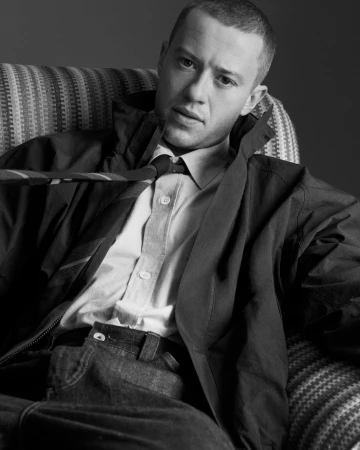 Portrait of a man in his 20s wearing an open collared shirt, tie, and jacket seated in a patterned armchair.