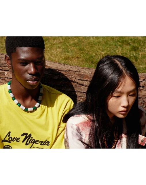 Portrait of an adult Black man and Asian woman wearing colorful shirts and beaded necklaces, sitting in an outdoor wooded environment.