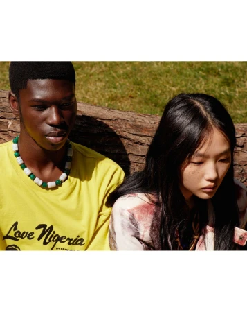Portrait of an adult Black man and Asian woman wearing colorful shirts and beaded necklaces, sitting in an outdoor wooded environment.