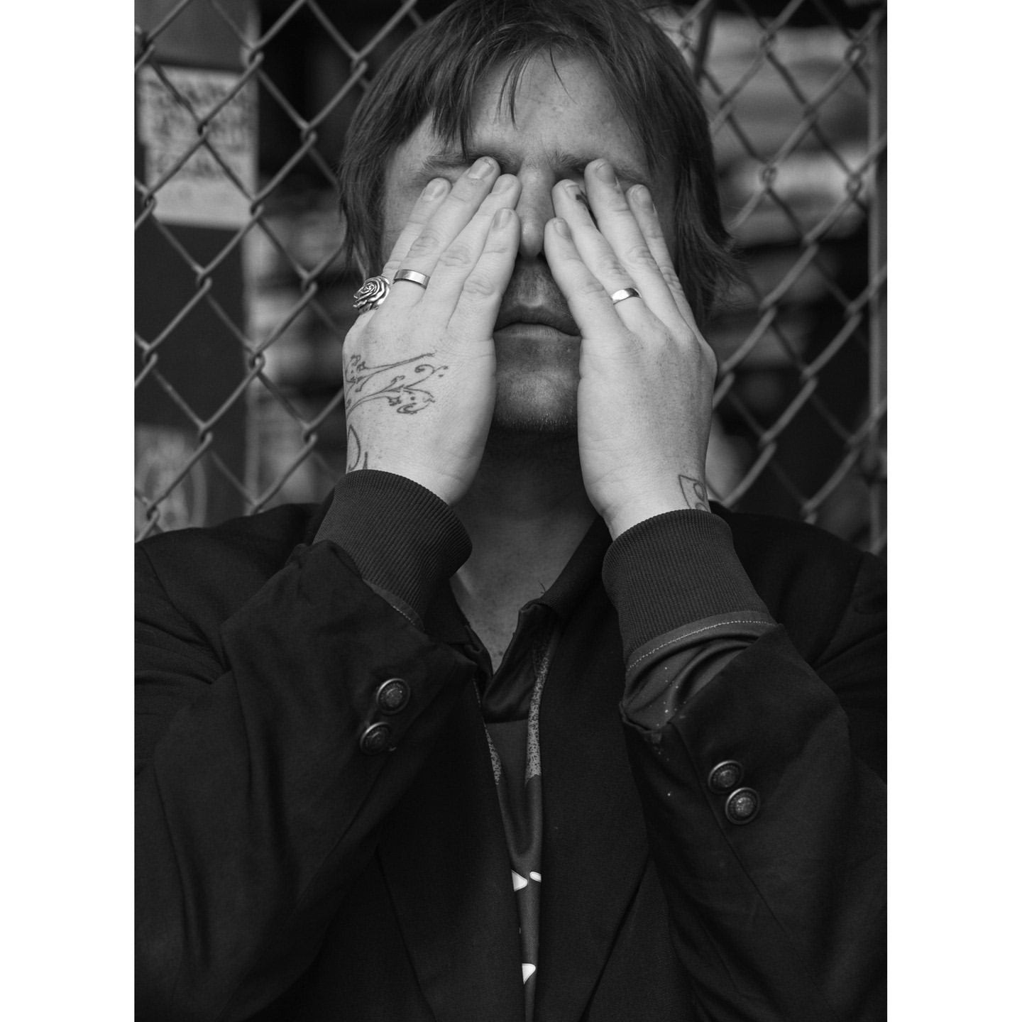 Monochrome portrait of an adult male covering his face with hands, featuring hand tattoos and rings, against a chain-link fence backdrop.