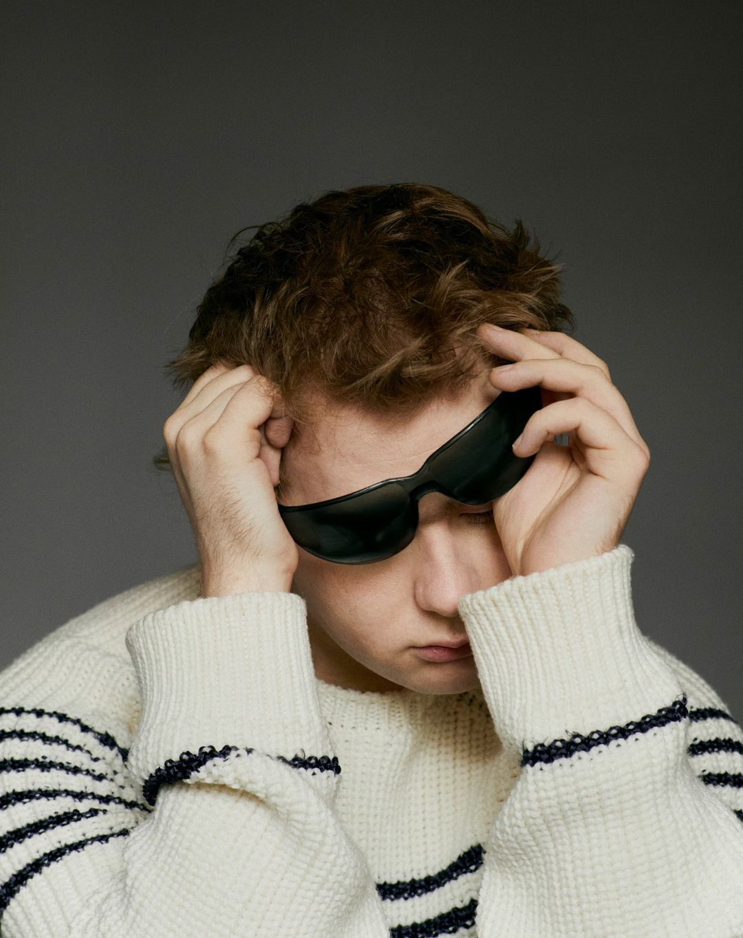 Young man with tousled hair and sunglasses in an eye-level portrait. He wears a striped sweater against a plain gray backdrop.