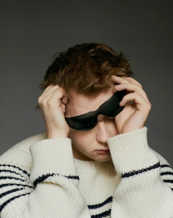 Young man with tousled hair and sunglasses in an eye-level portrait. He wears a striped sweater against a plain gray backdrop.