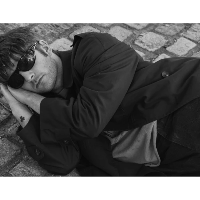Monochrome portrait of a man in sunglasses and coat lying on stone ground with hands near his face in an outdoor environment.