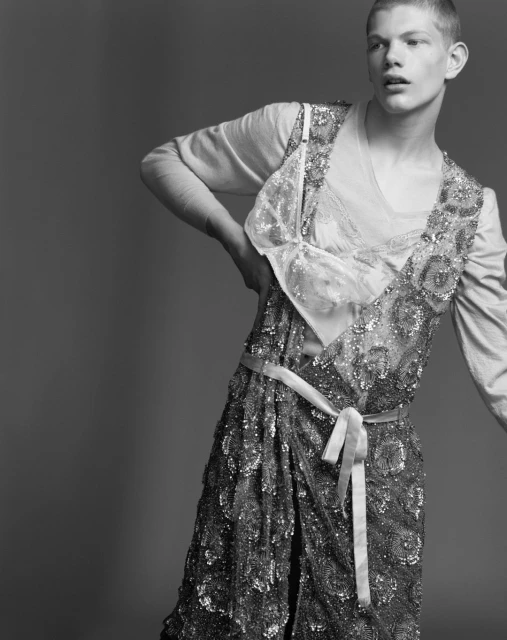 Monochrome fashion shot of a young adult posing in an embellished dress with an attached bra, a light-colored shirt, and a sash.