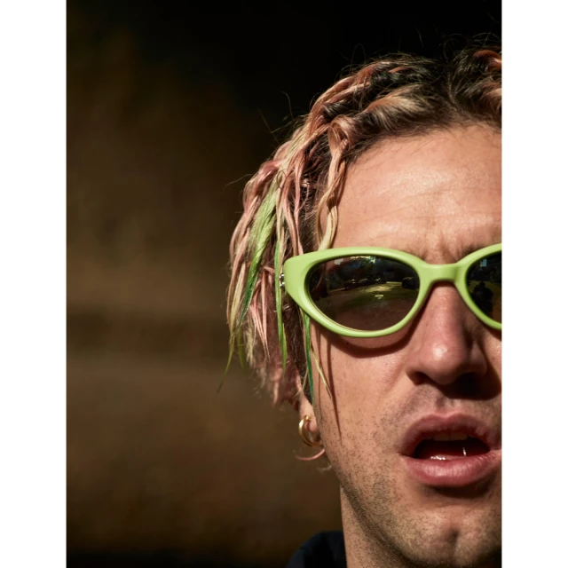 Portrait of a young adult man with dyed braided hair and lime green sunglasses in a close-up shot with a blurred background.
