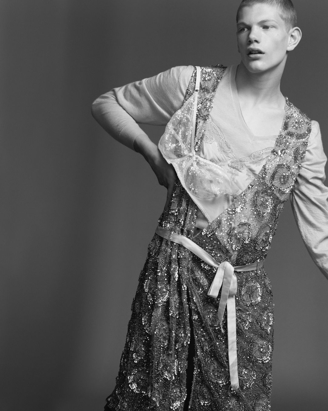 Black and white portrait of a young adult male model posing in a sequined dress with a ribbon belt, against a grey seamless backdrop.