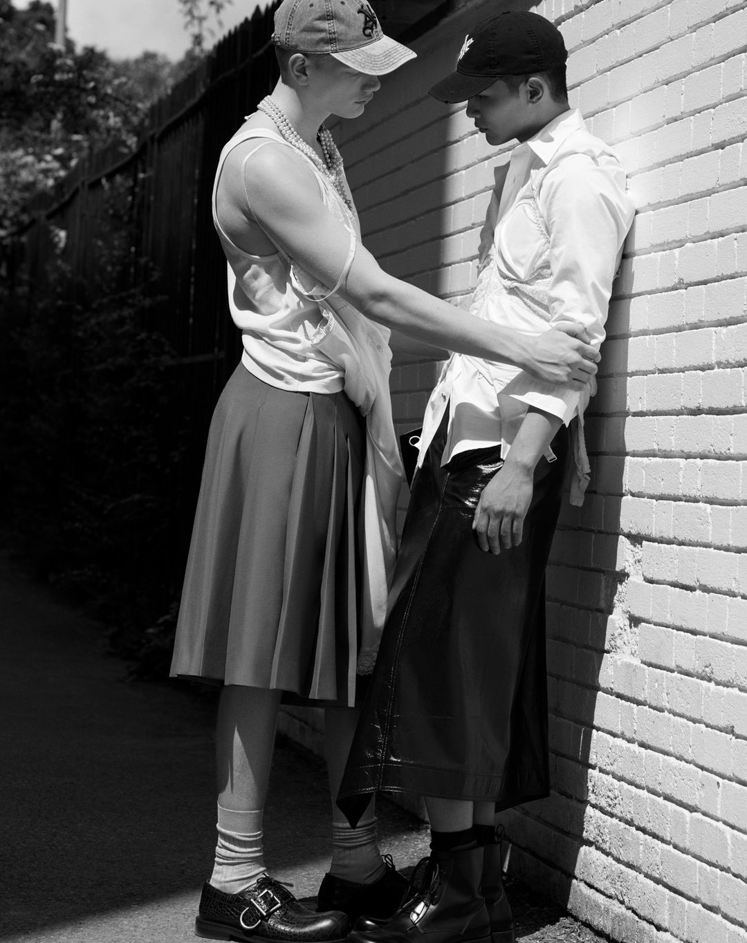 Two young men, one wearing a skirt and cap, lean against a brick wall in an urban alley in a black and white fashion shot.
