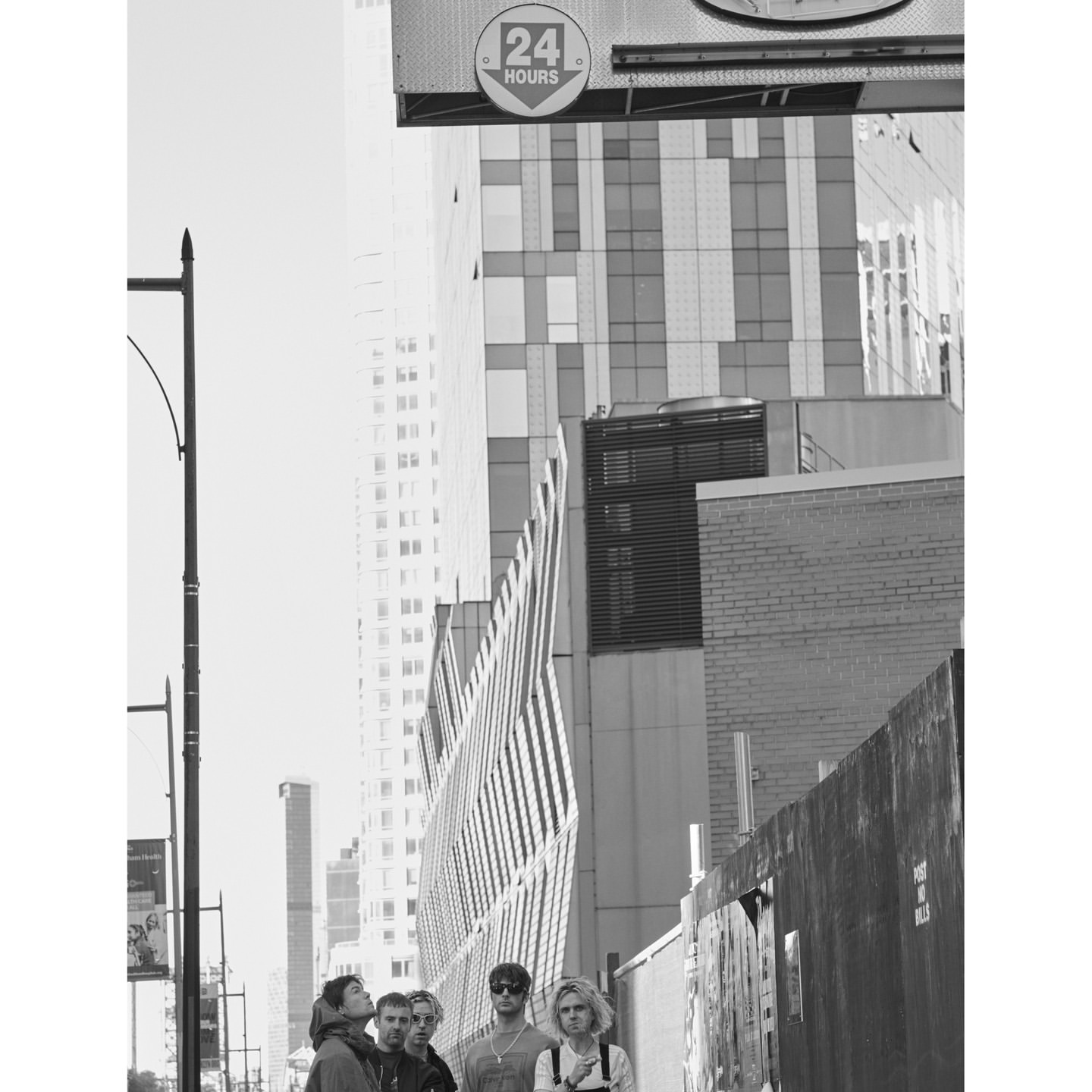 Group of young adults in city setting, monochromatic image features five people standing on a sidewalk with tall buildings in the background.