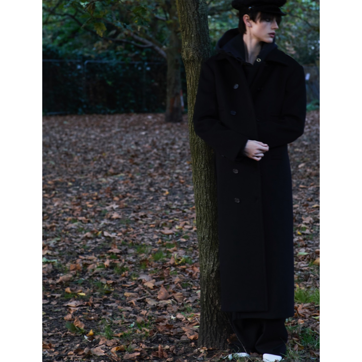 Full shot of a young adult male leaning against a tree in a park setting, wearing a black coat, cap, and white shoes in daylight.