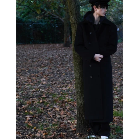 Full shot of a young adult male leaning against a tree in a park setting, wearing a black coat, cap, and white shoes in daylight.