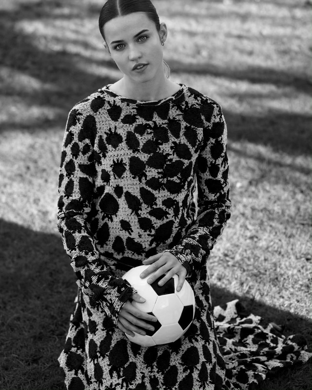 Monochrome portrait of a woman in a patterned dress kneeling on grass, holding a soccer ball with both hands, and looking at camera.
