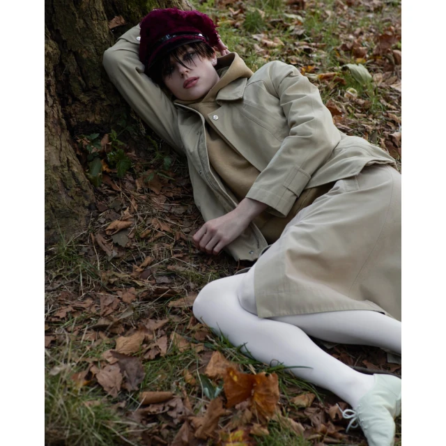 Young adult in beige jacket and skirt lies on forest floor by a tree, wearing a maroon hat and white tights, photographed in natural light.