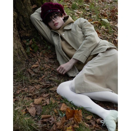 Young adult in beige jacket and skirt lies on forest floor by a tree, wearing a maroon hat and white tights, photographed in natural light.