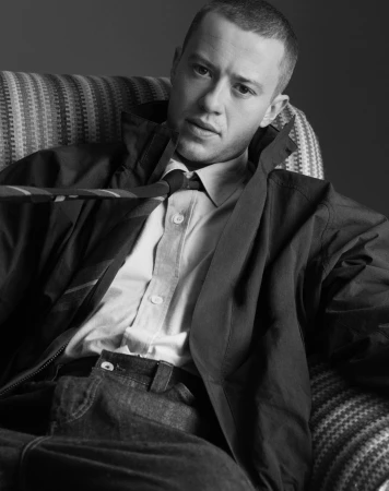 Portrait of a man in a collared shirt, jacket, and tie seated on a patterned sofa in a monochrome composition