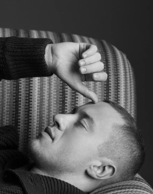 Black and white studio shot of an adult male lying down with a hand hovering above him with a thumbs-down gesture.