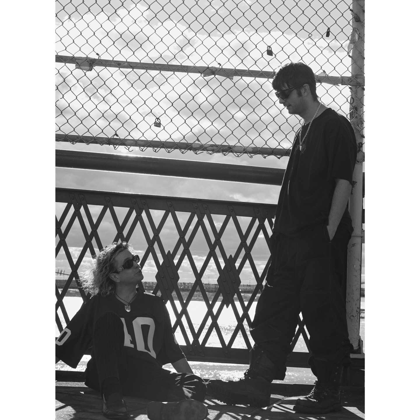 Two men in casual wear pose outdoors near a chain-link fence and metal structure on an overcast day, captured in black and white photography.