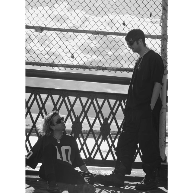 Two men in casual wear pose outdoors near a chain-link fence and metal structure on an overcast day, captured in black and white photography.