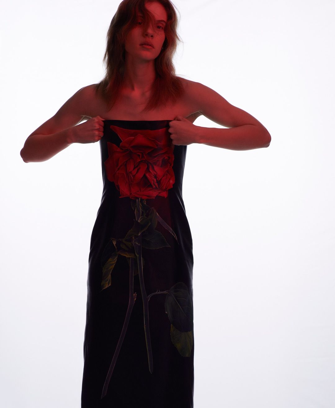Model poses in a dark dress with a red rose design against a bright white background in a fashion photography studio shot.