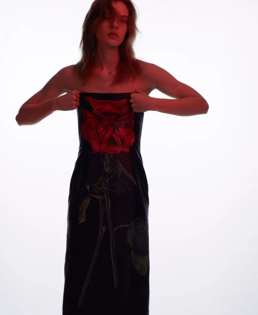 Model poses in a dark dress with a red rose design against a bright white background in a fashion photography studio shot.