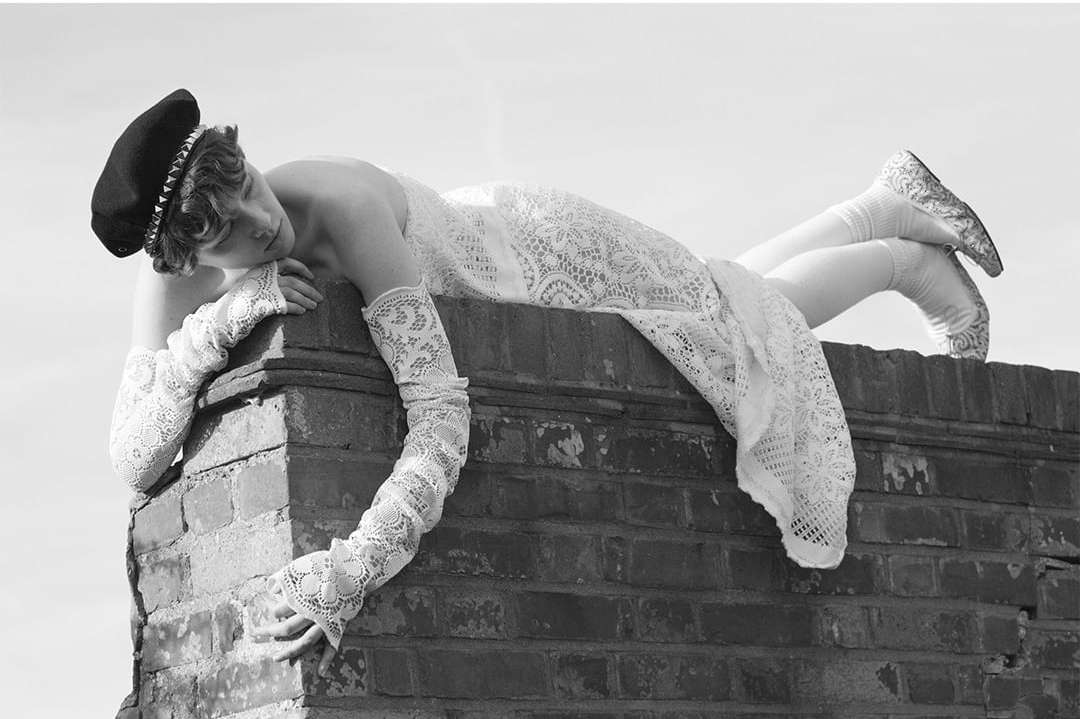 Monochrome portrait of a woman wearing a beret and a white lace dress and gloves, lying on a brick structure against a clear sky.