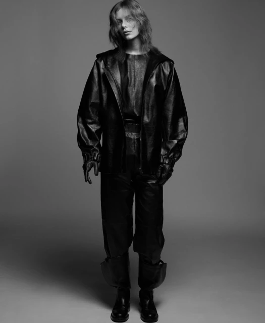 Black and white studio shot of a woman posing in a full-length leather outfit, including a jacket, pants, and gloves, against a grey backdrop.