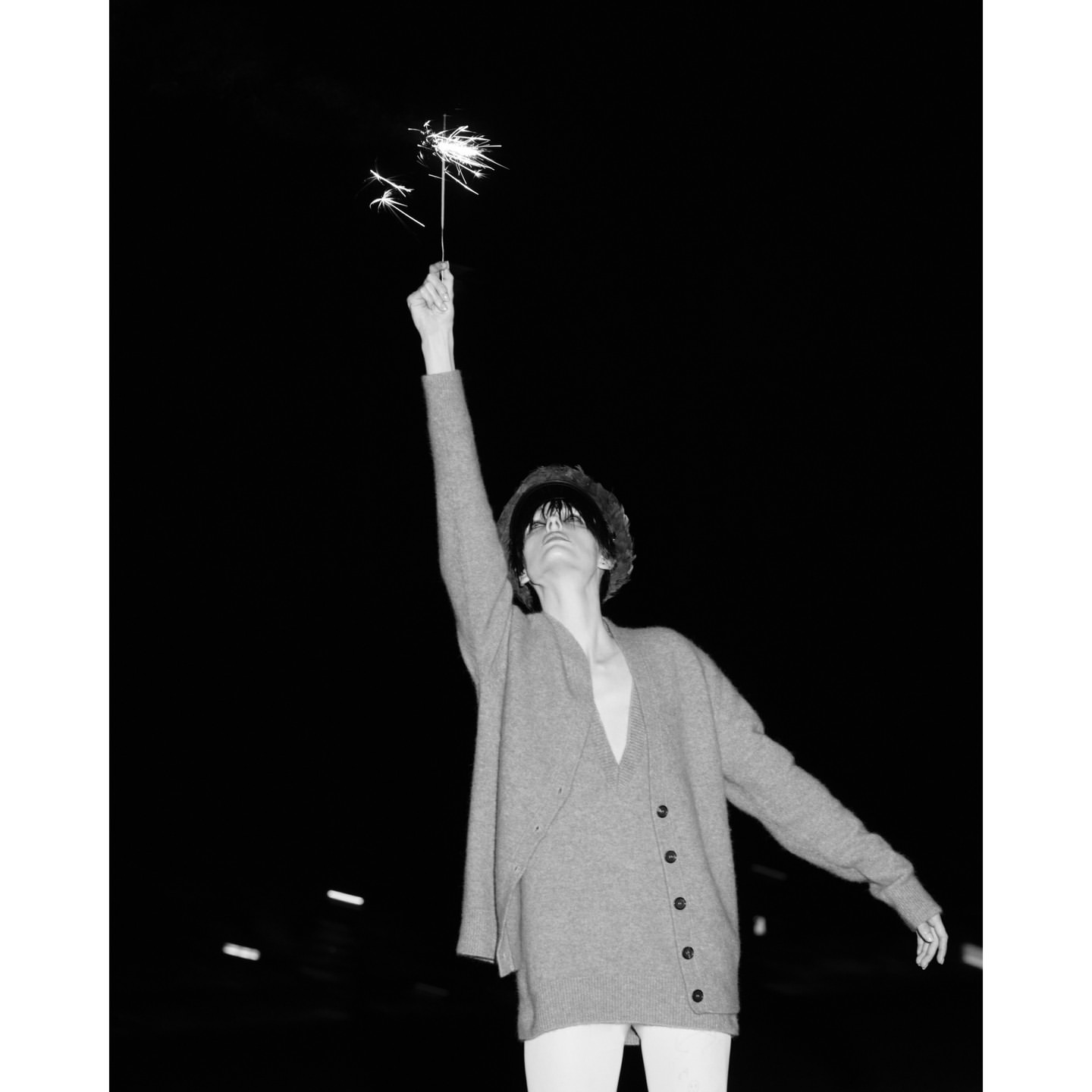 Monochrome portrait of a woman holding sparklers against a dark night sky, wearing a cardigan and hat, with visible dark hair and bangs.
