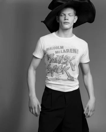 Monochrome portrait of a slender adult male with eyes closed, wearing a graphic tee, wide-leg pants, and wide-brimmed hat in a studio.