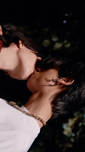 Close-up vertical shot of two people kissing outdoors. One is wearing large silver earrings and a beaded necklace.