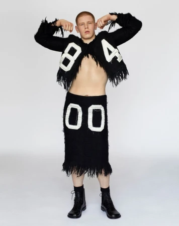 Young man with short red hair flexing in black fringed two-piece outfit with white numbers 84 and 00 against white background.