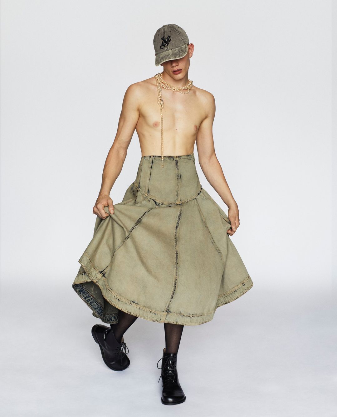 Young adult poses in a cap, beaded necklace, olive green skirt with black tights and boots against a clean, white backdrop in studio lighting.