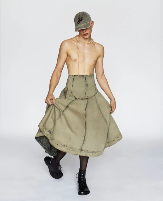 Young adult poses in a cap, beaded necklace, olive green skirt with black tights and boots against a clean, white backdrop in studio lighting.