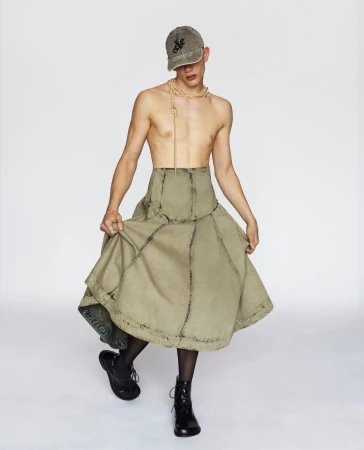 Young adult poses in a cap, beaded necklace, olive green skirt with black tights and boots against a clean, white backdrop in studio lighting.