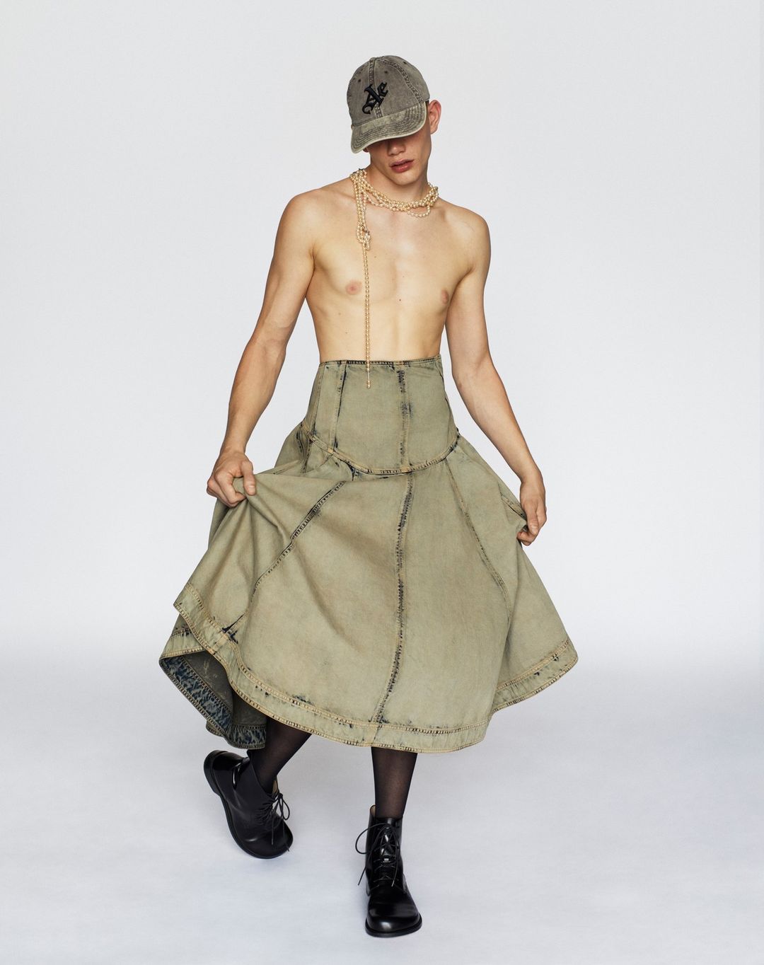 Fashion photo of a shirtless adult male model wearing a khaki baseball cap, tan necklace, large tan skirt, and black boots in studio.