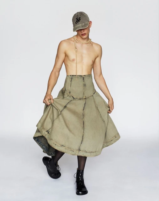 Fashion photo of a shirtless adult male model wearing a khaki baseball cap, tan necklace, large tan skirt, and black boots in studio.