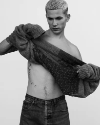 Man with short blonde hair taking off a dark knitted sweater against a plain white background