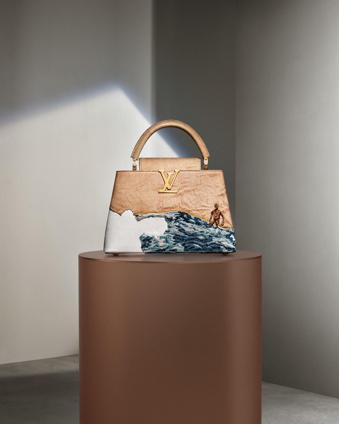 Fashion still life: A beige Louis Vuitton Capucines bag sits on a brown plinth against a pale grey wall, with a small figurine.