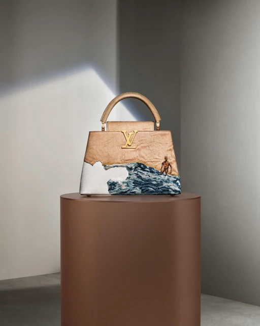 Fashion still life: A beige Louis Vuitton Capucines bag sits on a brown plinth against a pale grey wall, with a small figurine.