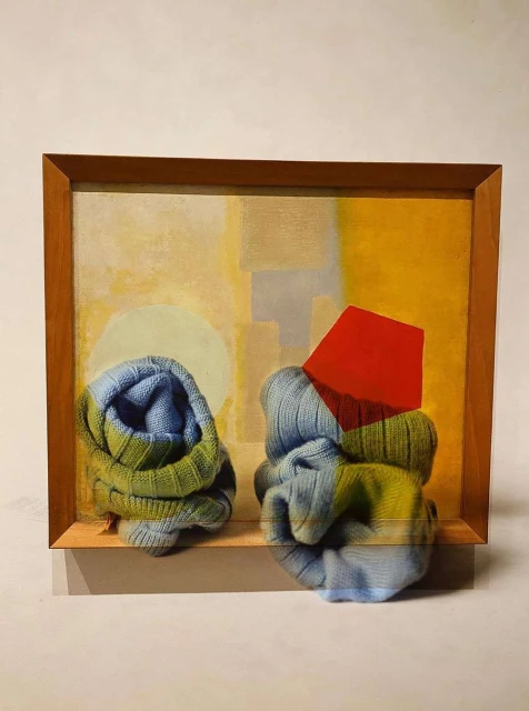 Still life showcases art installation with folded blue and green knit fabric and a red geometric shape against a yellow background inside a brown frame.