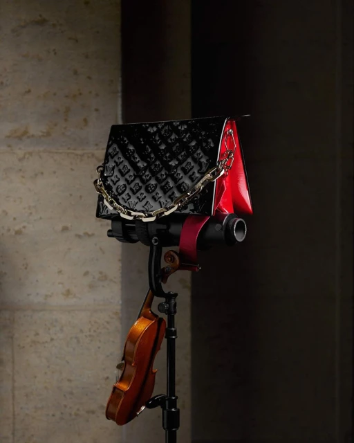 A black, patent leather purse with a gold chain is displayed on a black stand next to a violin against a tan wall backdrop.