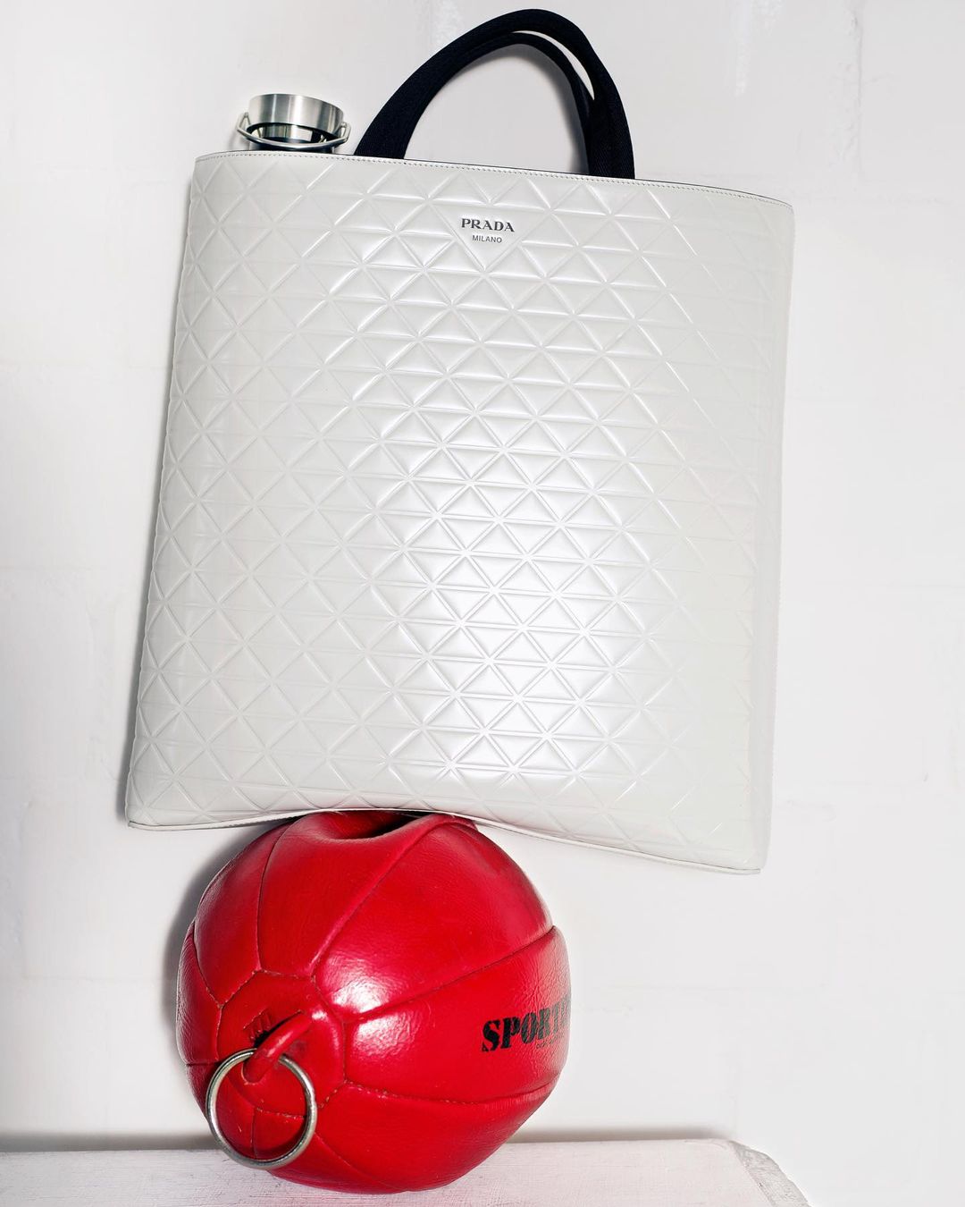 Ivory-colored Prada tote bag with black straps rests atop a red sports ball with silver rings in a minimalist setting.