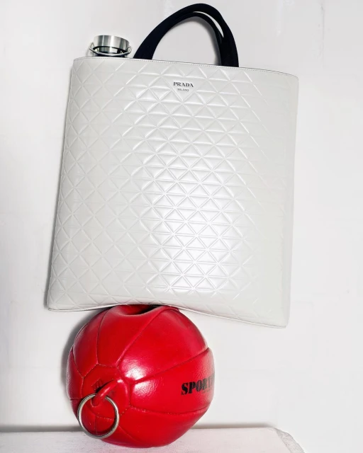 Ivory-colored Prada tote bag with black straps rests atop a red sports ball with silver rings in a minimalist setting.