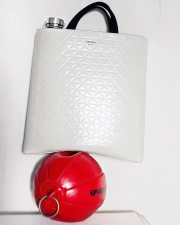 Ivory-colored Prada tote bag with black straps rests atop a red sports ball with silver rings in a minimalist setting.