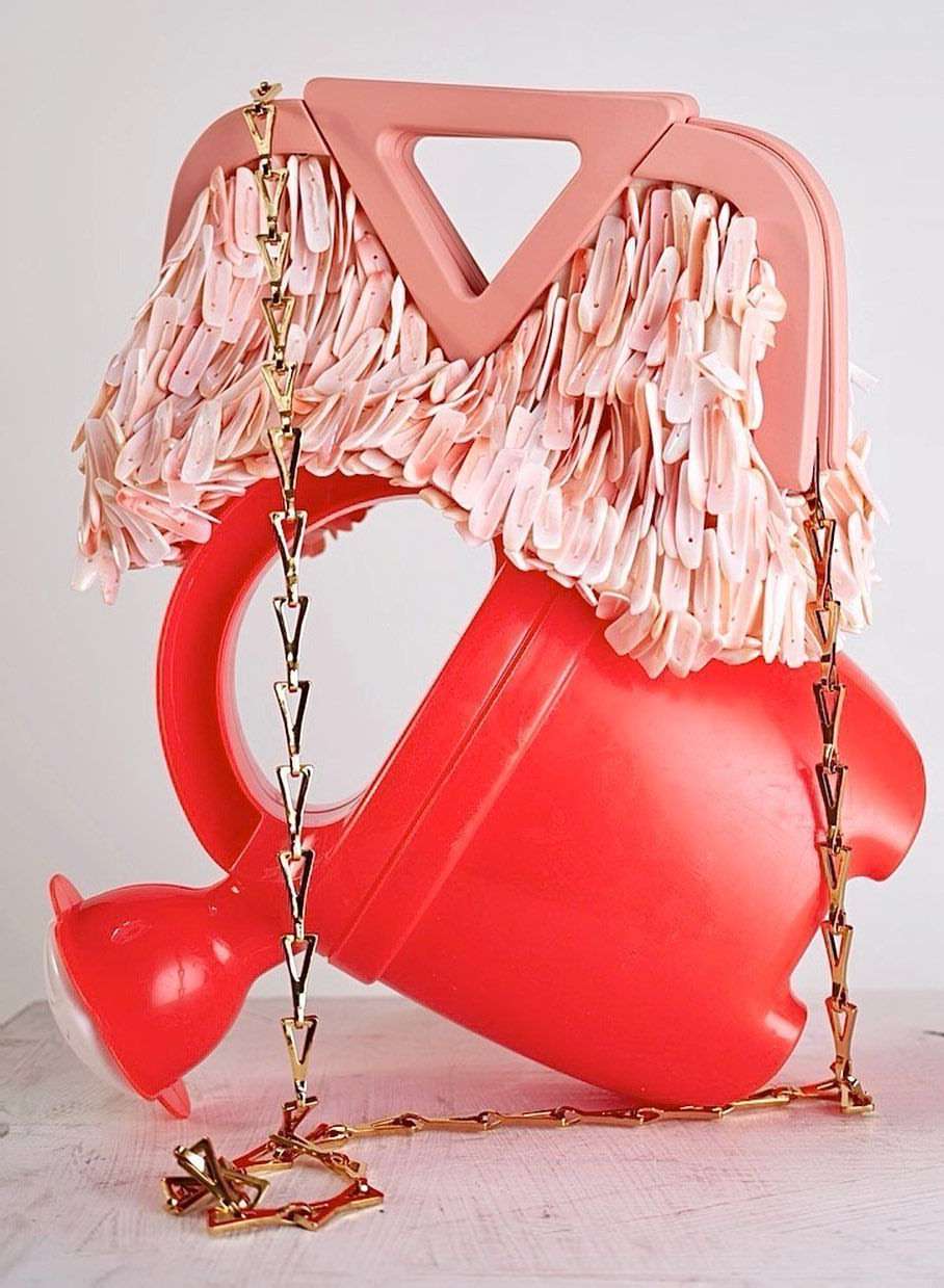 Artistic still life featuring a sculptural handbag made from a watering can, adorned with fringe and a gold chain, against a neutral backdrop.