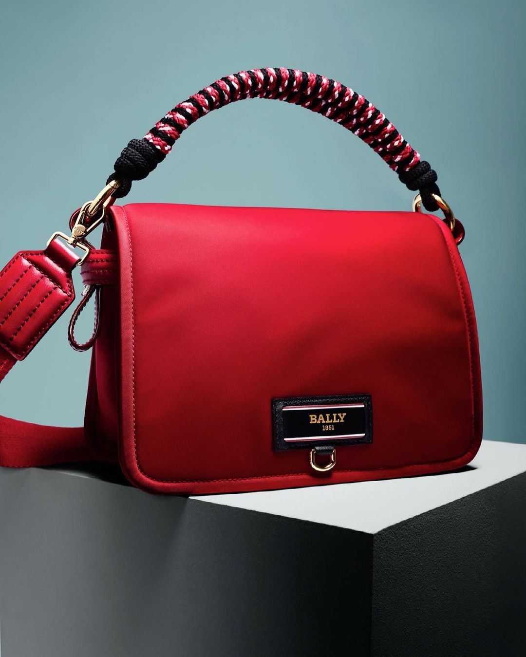 Studio shot of a red nylon Bally handbag with a braided handle, gold hardware, and a black-and-gold logo plate. Fashion product photo.
