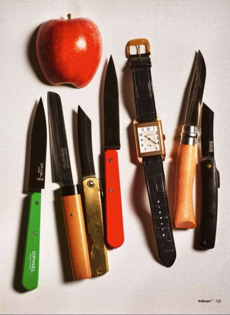Still life of a red apple, a gold watch, and an arrangement of pocket knives with green, gold, and red handles on a white surface.