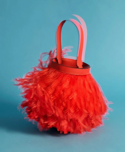 Close-up studio shot of a vibrant red feathered handbag with orange straps against a turquoise background, emphasizing texture and color.