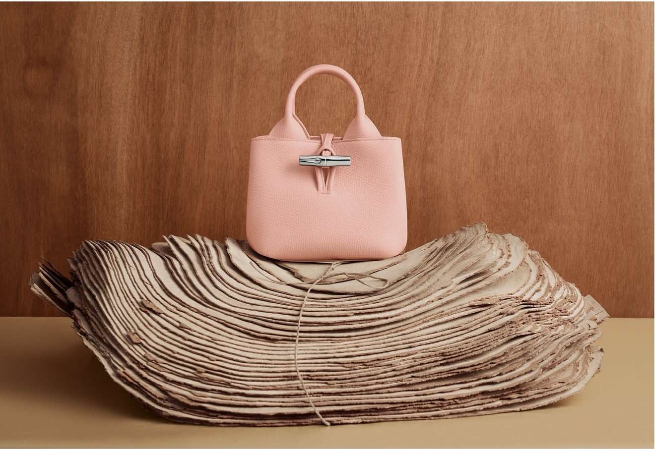Stylish pink leather handbag on sculpted paper against a wood backdrop. Fashion product shot with luxury feel, studio lighting, and clean style.