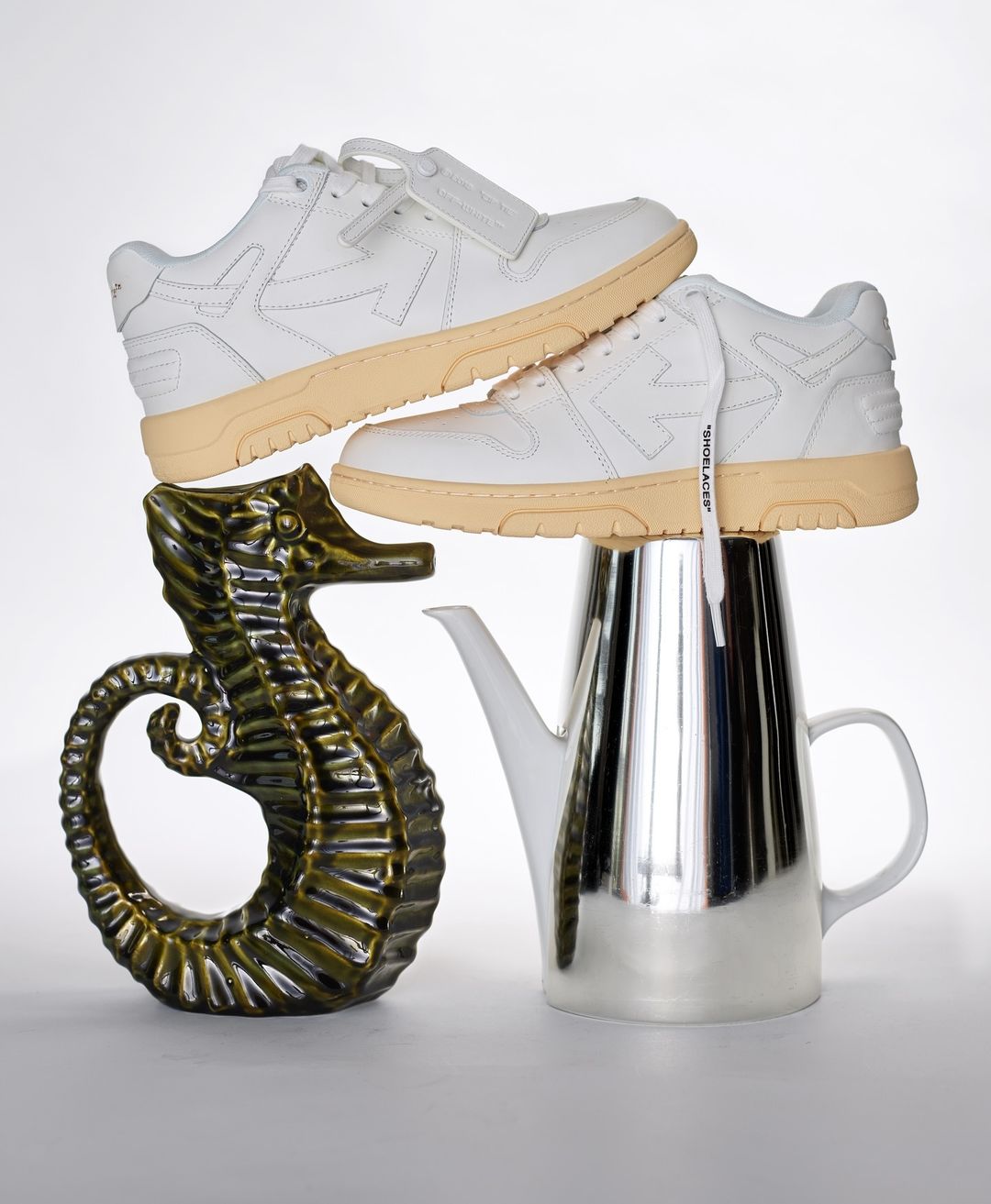 Elevated view of stacked white sneakers, silver teapot, and green seahorse figurine against a white backdrop in a studio setting.